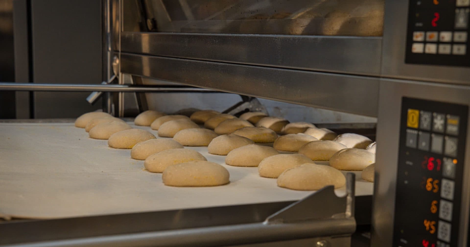 Forno industriale a più piani in acciaio inox con impasti infarinati su un nastro di carico e pannelli di controllo digitali